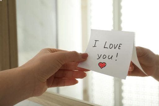 Close-up of a woman’s hand holding a sticky note stuck to a mirror with “I love you” written. 