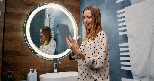 Woman smiling and looking at her tablet while holding a toothbrush in the bathroom.