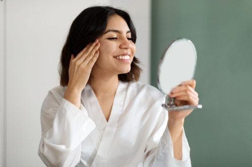 Woman wearing a white robe smiling and touching her face in front of a mirror.